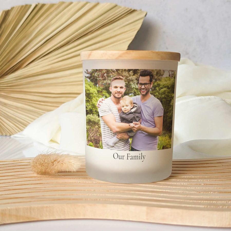 The Personalized Picture Candle with Custom Photo by Little Piece Candle Co. sits on a wooden slat surface, featuring a photo of two men and a baby, labeled "Our Family," with dried plants decorating the background.