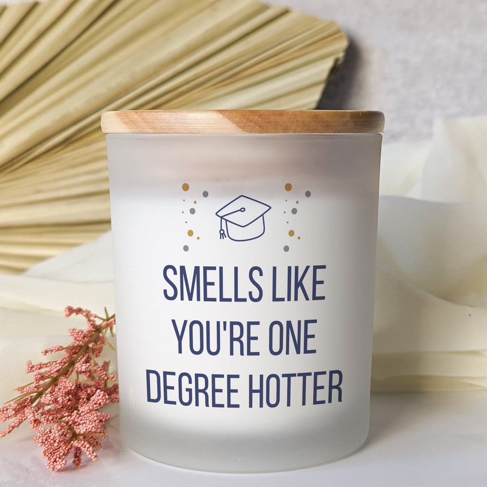 Frosted candle reading “Smells Like You’re One Degree Hotter,” with a graduation cap, confetti dots and a wooden lid.