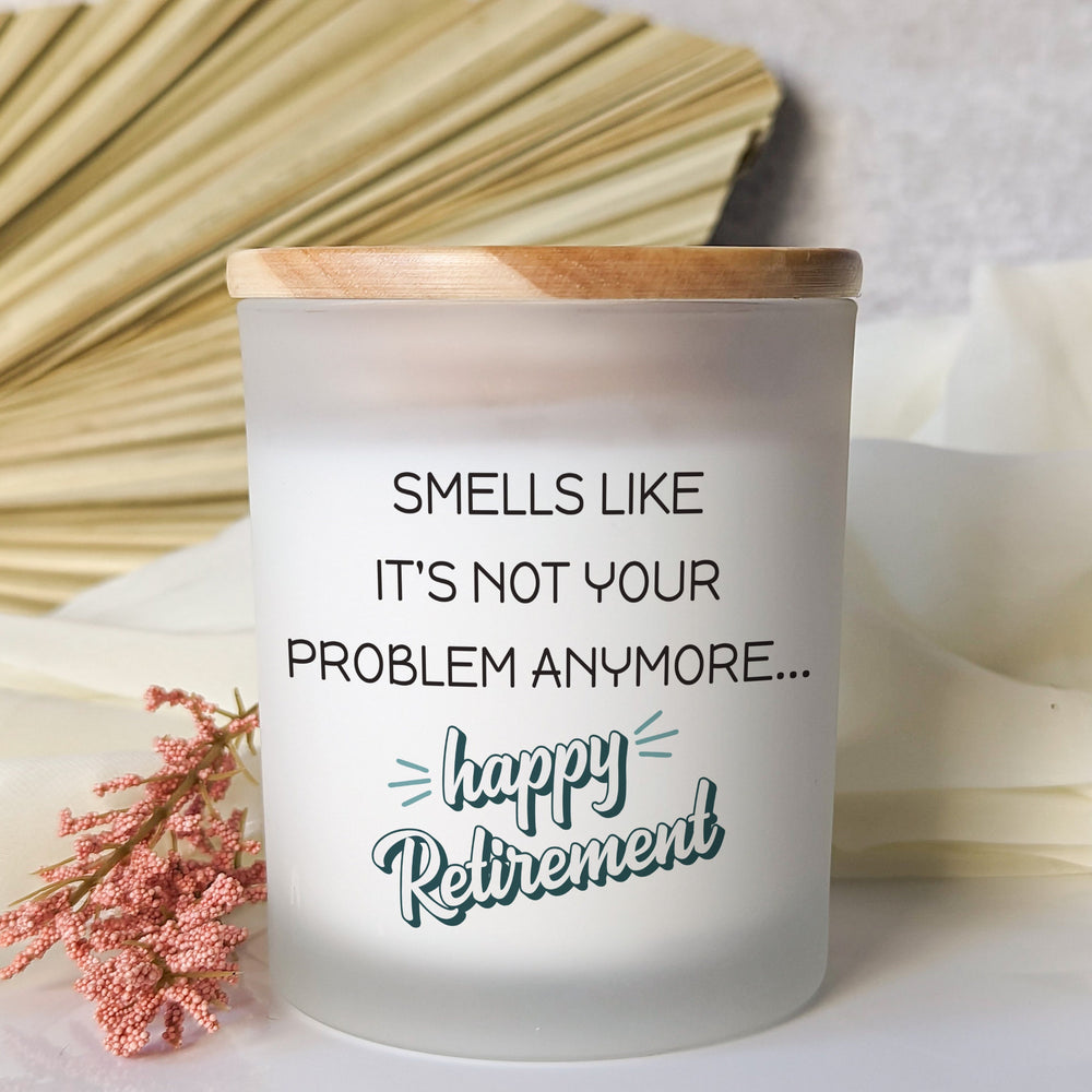A frosted glass candle jar from Little Piece Candle Co. with a wooden lid, labeled “Smells like it’s not your problem anymore... happy retirement.”