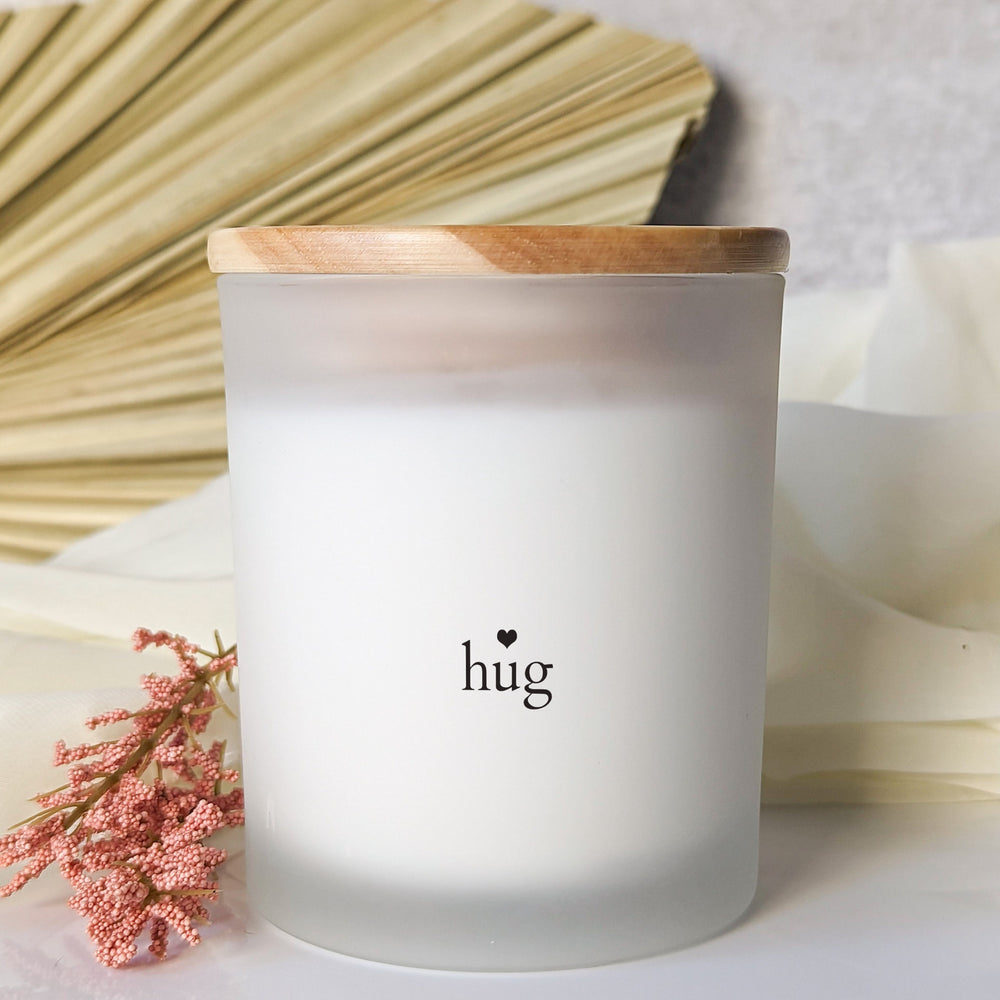 Frosted candle with small “hug” lettering and a black heart, topped with a wooden lid beside pink dried flowers and a palm leaf.