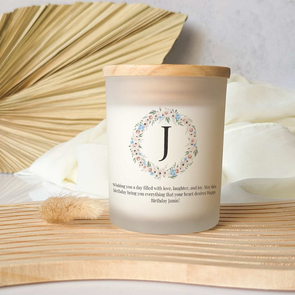 A Little Piece Candle Co. Personalized Scented Candle with Initial and Your Text in a frosted glass jar with a wooden lid, featuring a floral wreath, the letter “J,” and a custom birthday message printed on the front.