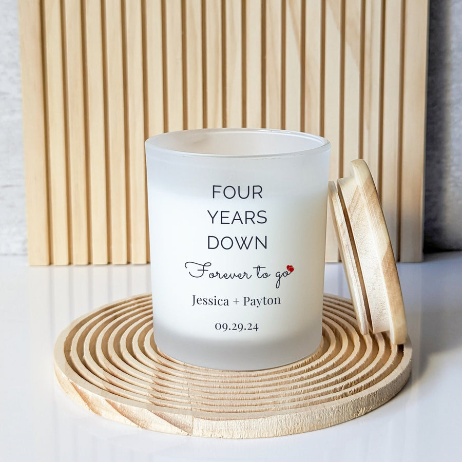 Frosted “Four Years Down, Forever to go” anniversary candle on a round grooved coaster, with its wooden lid leaning beside it.